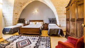 A bedroom with a large arched stone ceiling, a wooden bed, two smaller beds, a red chair, and a patterned rug. - Laurito Resort & Spa (Oria)