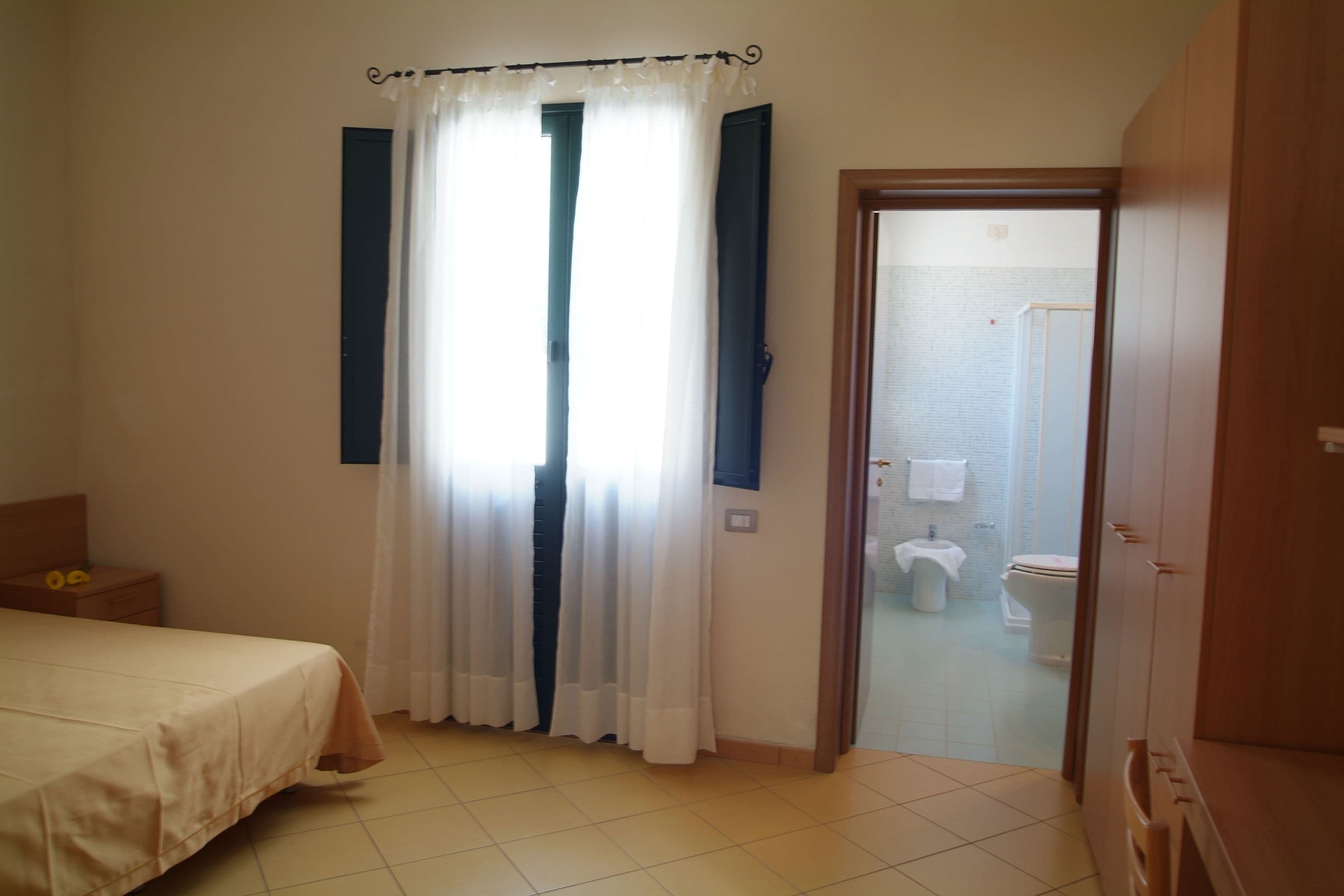 A bedroom with a bed, a window with curtains, a wooden wardrobe, and a bathroom visible through an open door.