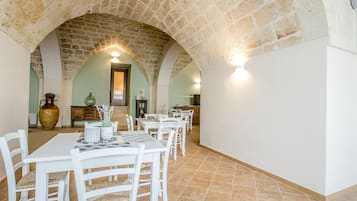 Un espace repas avec des tables et des chaises blanches, un plafond en forme d’arc en pierre et une cuisine en arrière-plan.