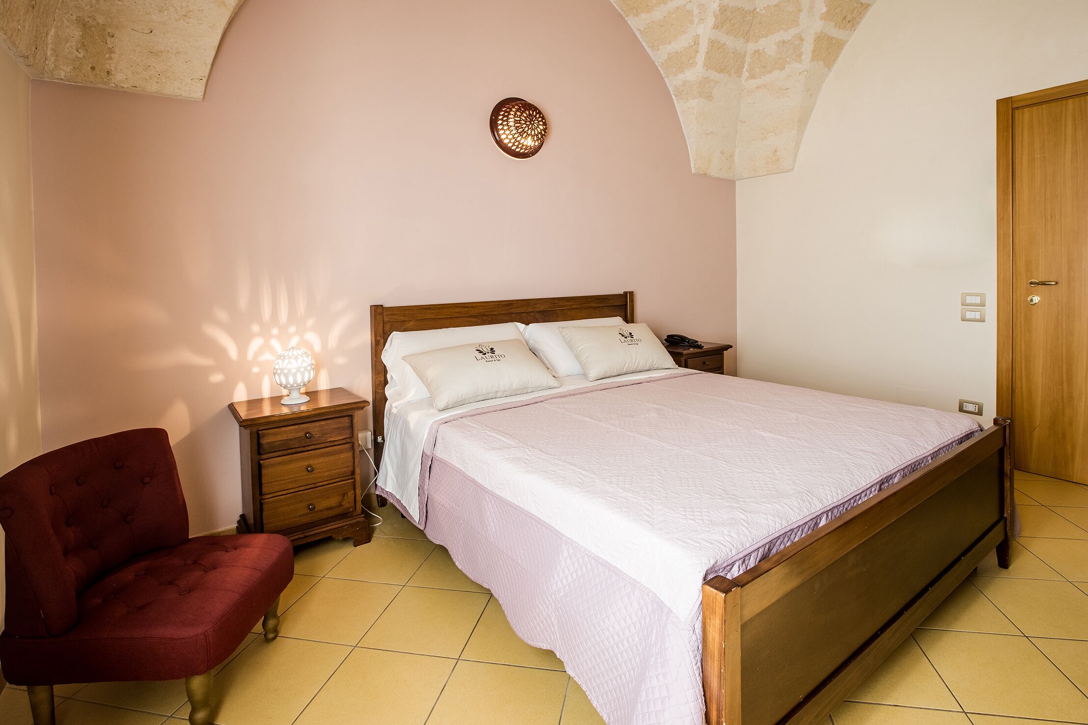 Quarto com cama de madeira, mesa de cabeceira, cadeira vermelha e luminária fixada na parede.