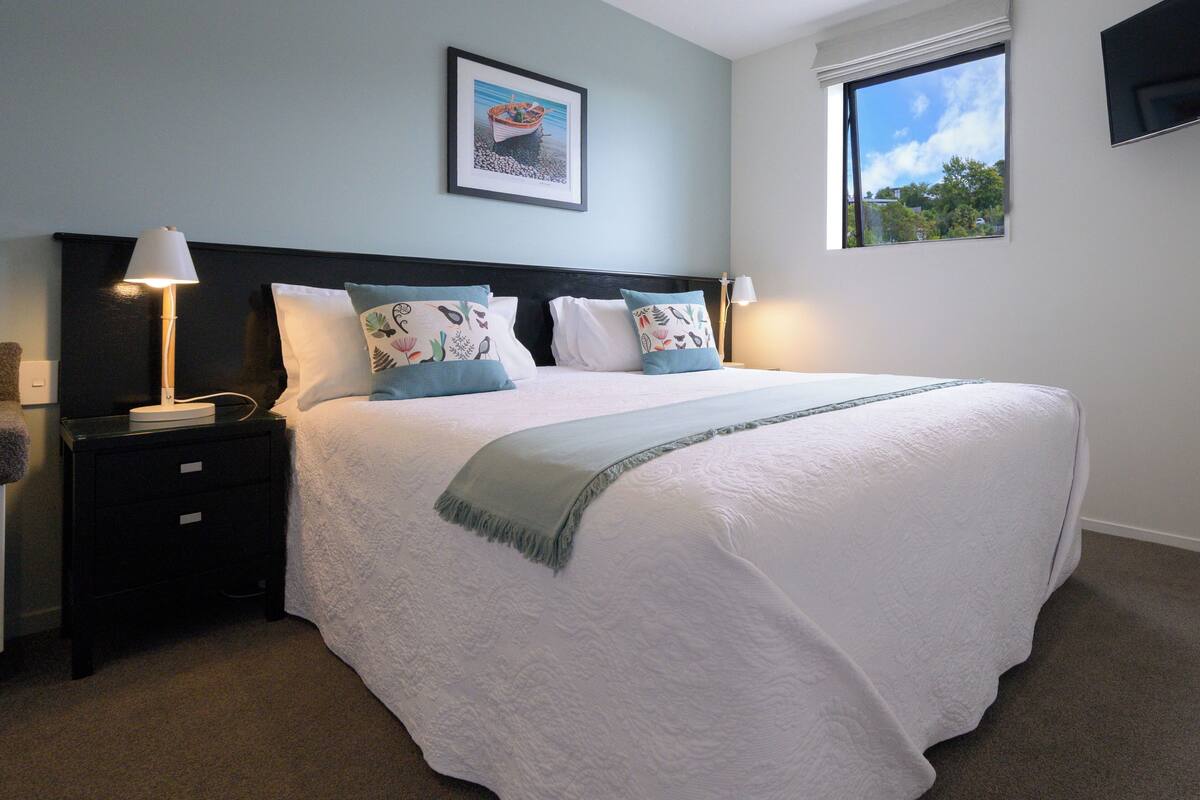 A neatly made bed with a white comforter and decorative pillows, a bedside lamp, and a framed picture on the wall.