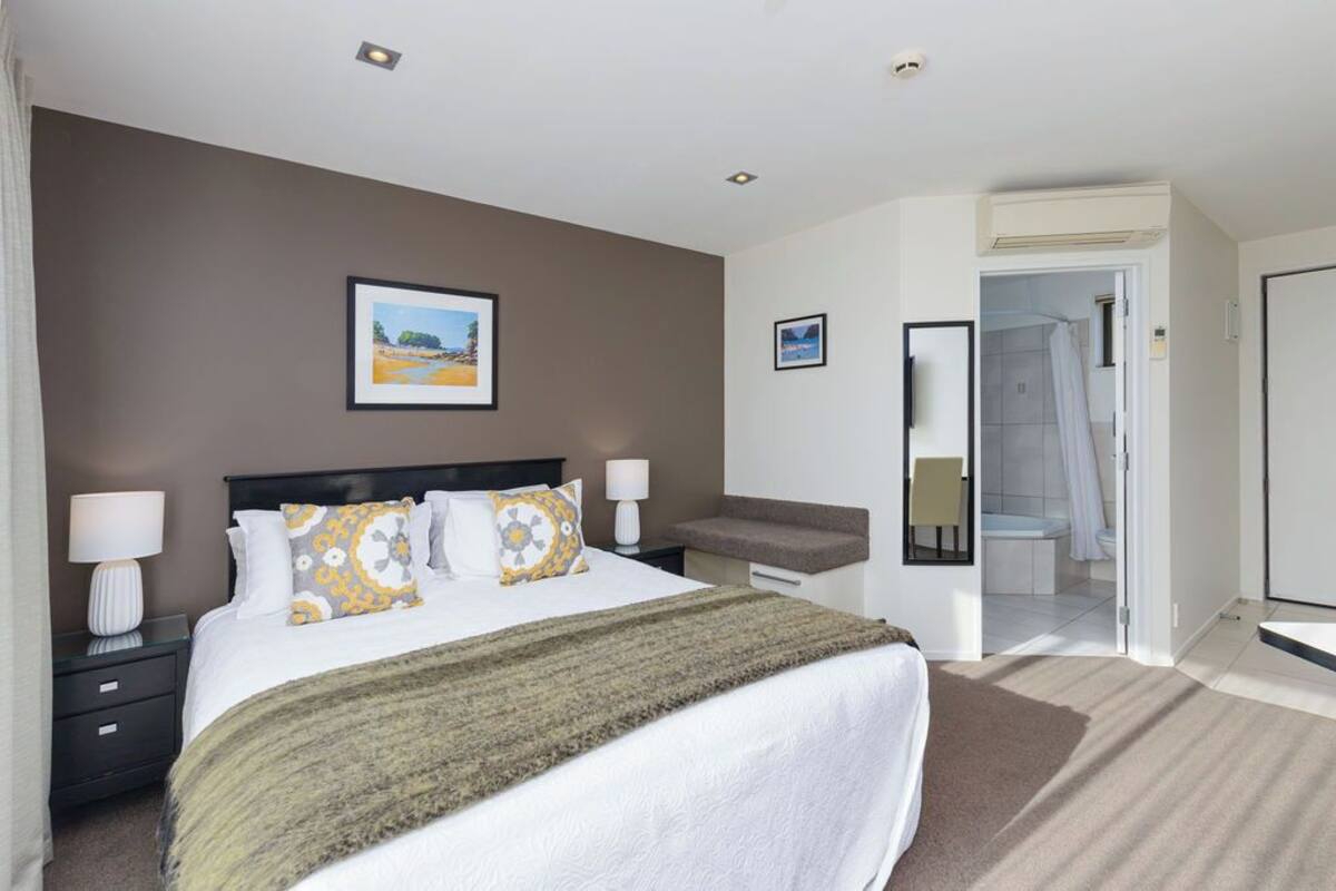 A hotel room with a large bed, bedside tables, a small desk, a chair, a shower area, and a door leading to another room.