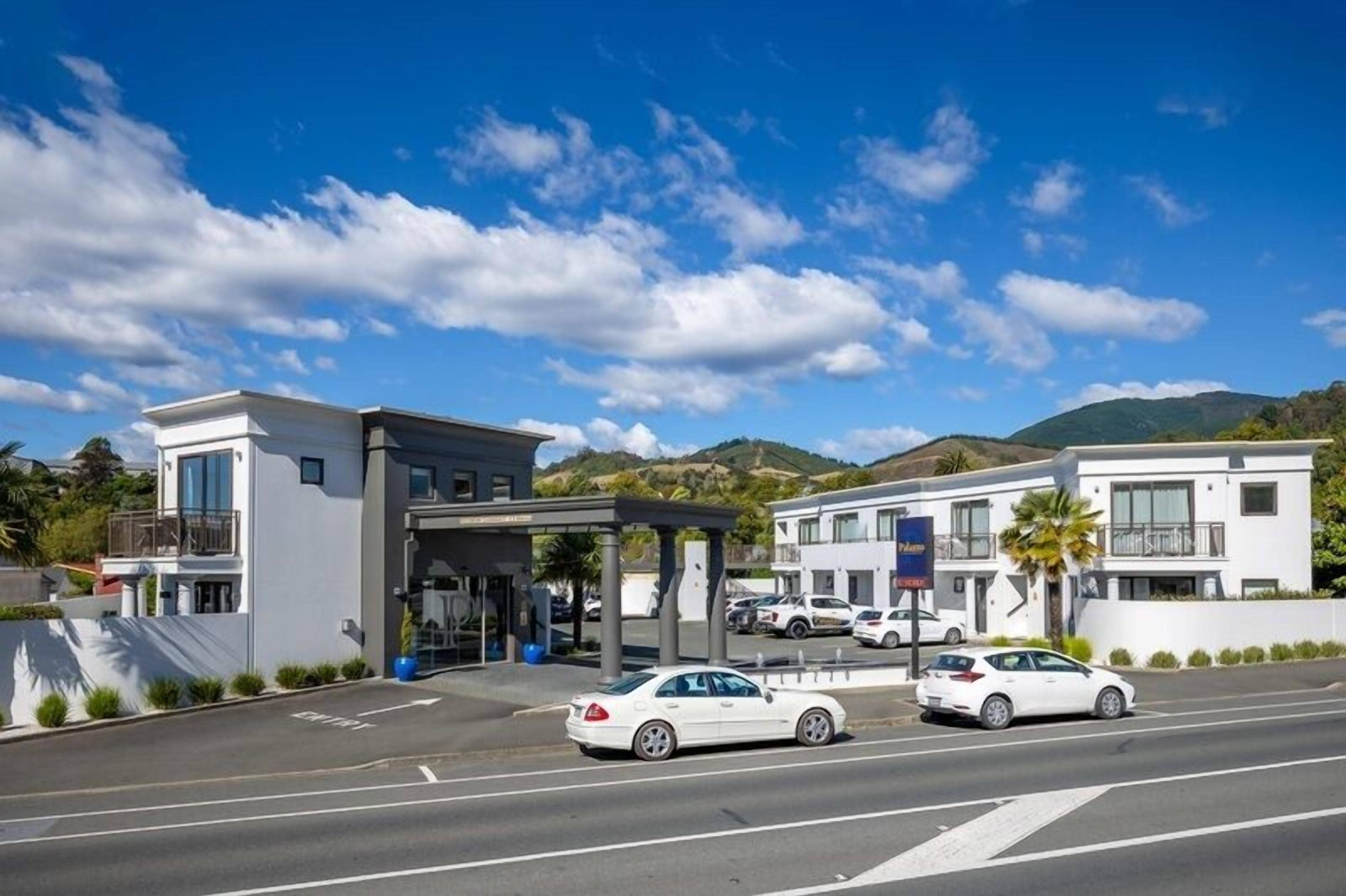 Un hotel moderno con un acceso cubierto, varios espacios de estacionamiento y vistas a las colinas al fondo.