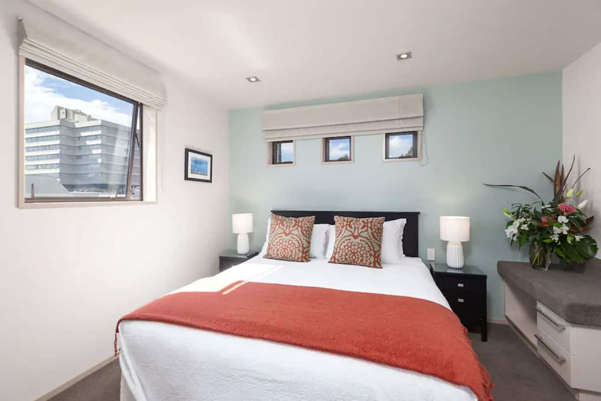 A hotel room with a large bed, two bedside tables with lamps, a window with a view of a building, and a small plant on the nightstand.