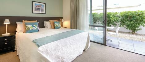 A bedroom with a large bed, a nightstand, a lamp, and a window with a view of greenery.
