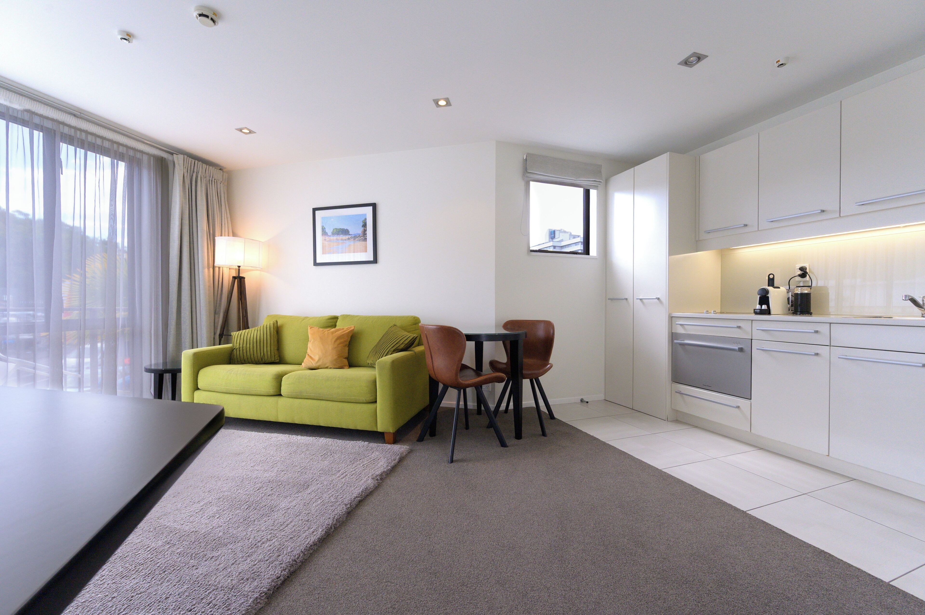 Una habitación de hotel moderna con un sofá verde, dos sillas, una mesita, una lámpara y una zona de cocina.