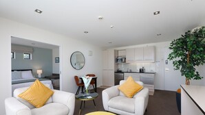 Ett modernt hotellrum med två vita fåtöljer, en gul pall, ett matbord med stolar, en köksdel och en stor grön växt.
