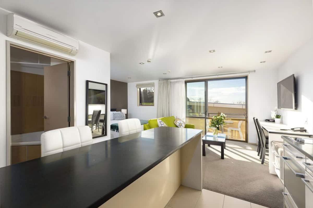 A modern hotel room with a large central dining table, a sofa, a TV, and a balcony with a view.