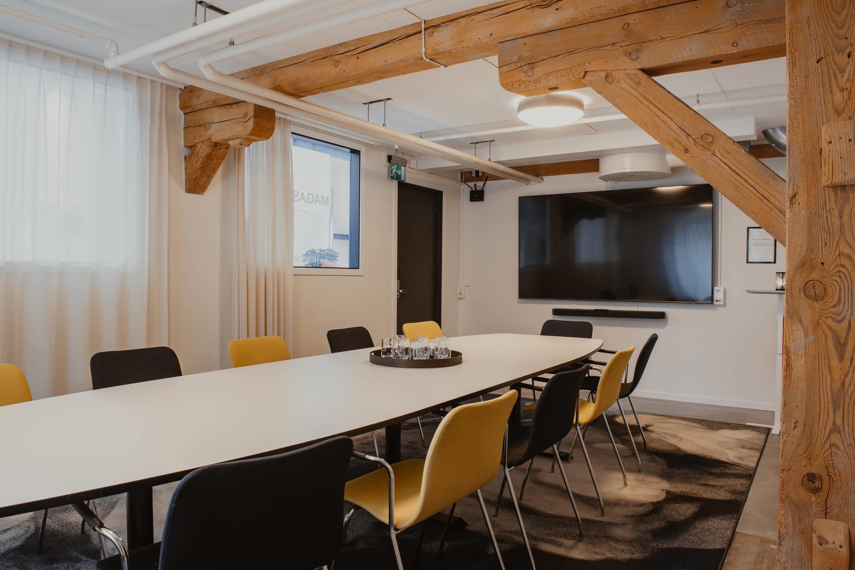 A conference room with a long table, yellow and black chairs, wooden beams, a large screen, and a window with curtains.