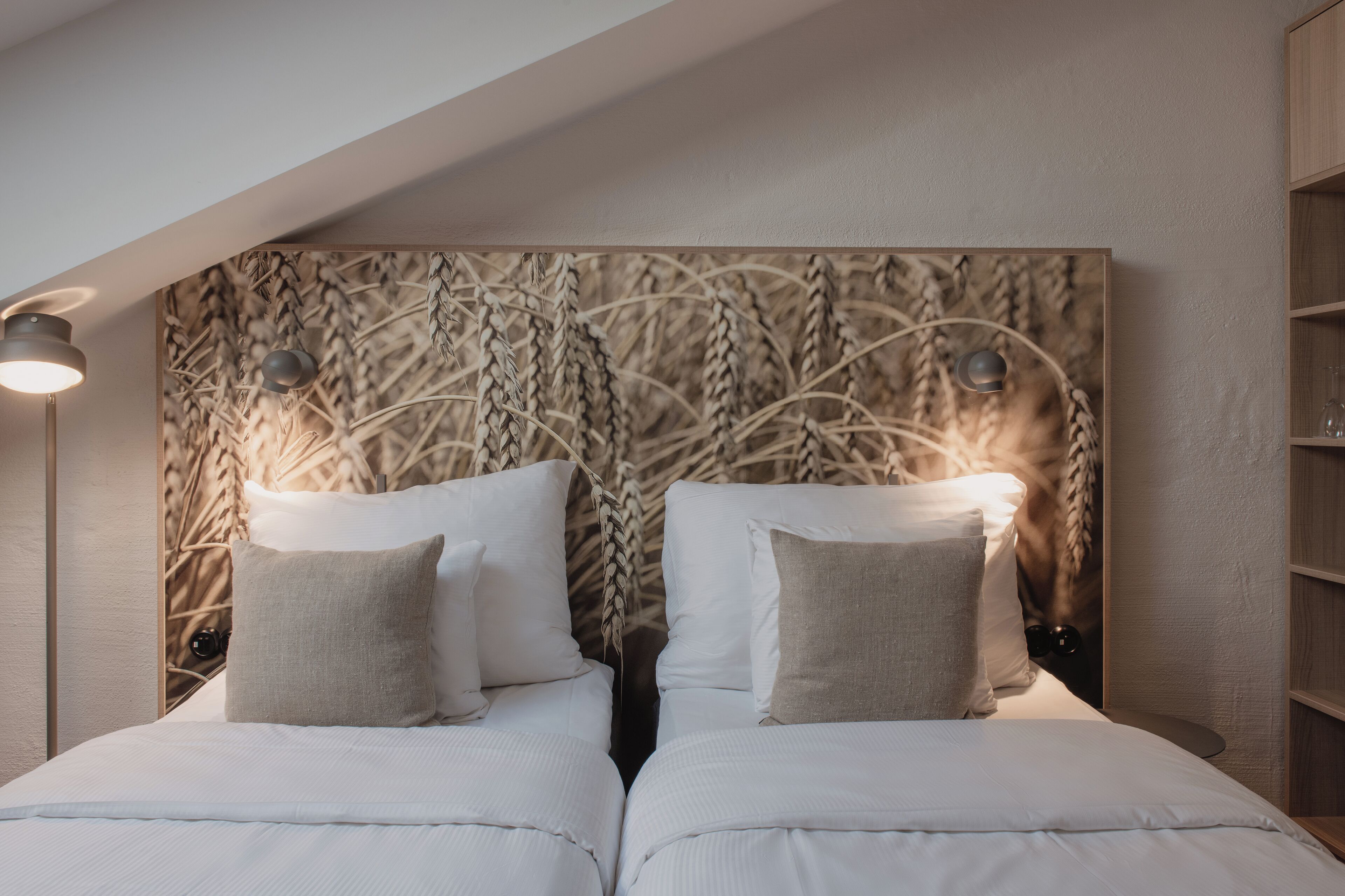 A bedroom with a headboard featuring a wheat pattern, two beds with white bedding, and a bedside lamp.