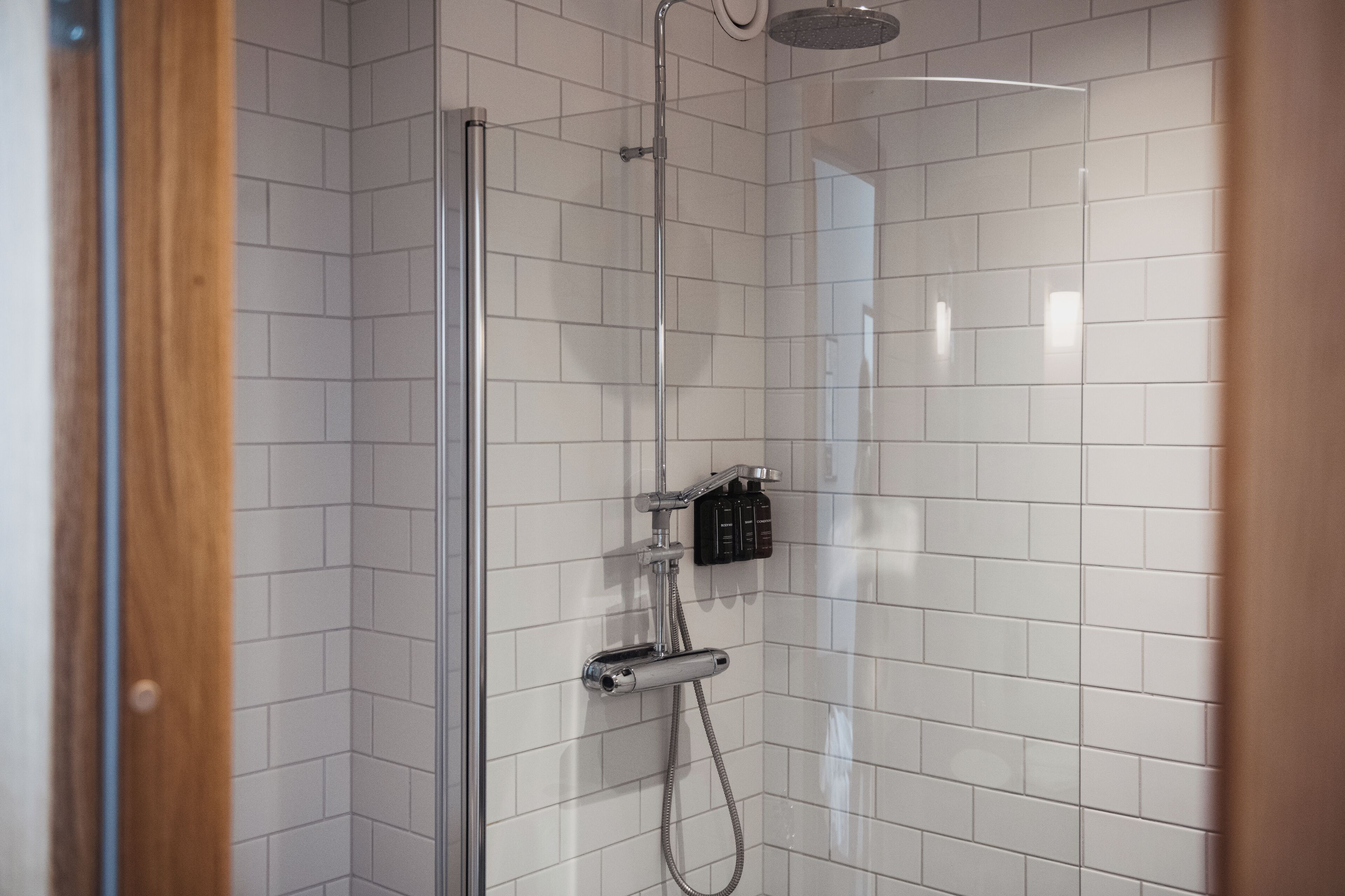 Un espace douche avec des murs recouverts de carreaux blancs, une porte en verre et une pomme de douche à main.