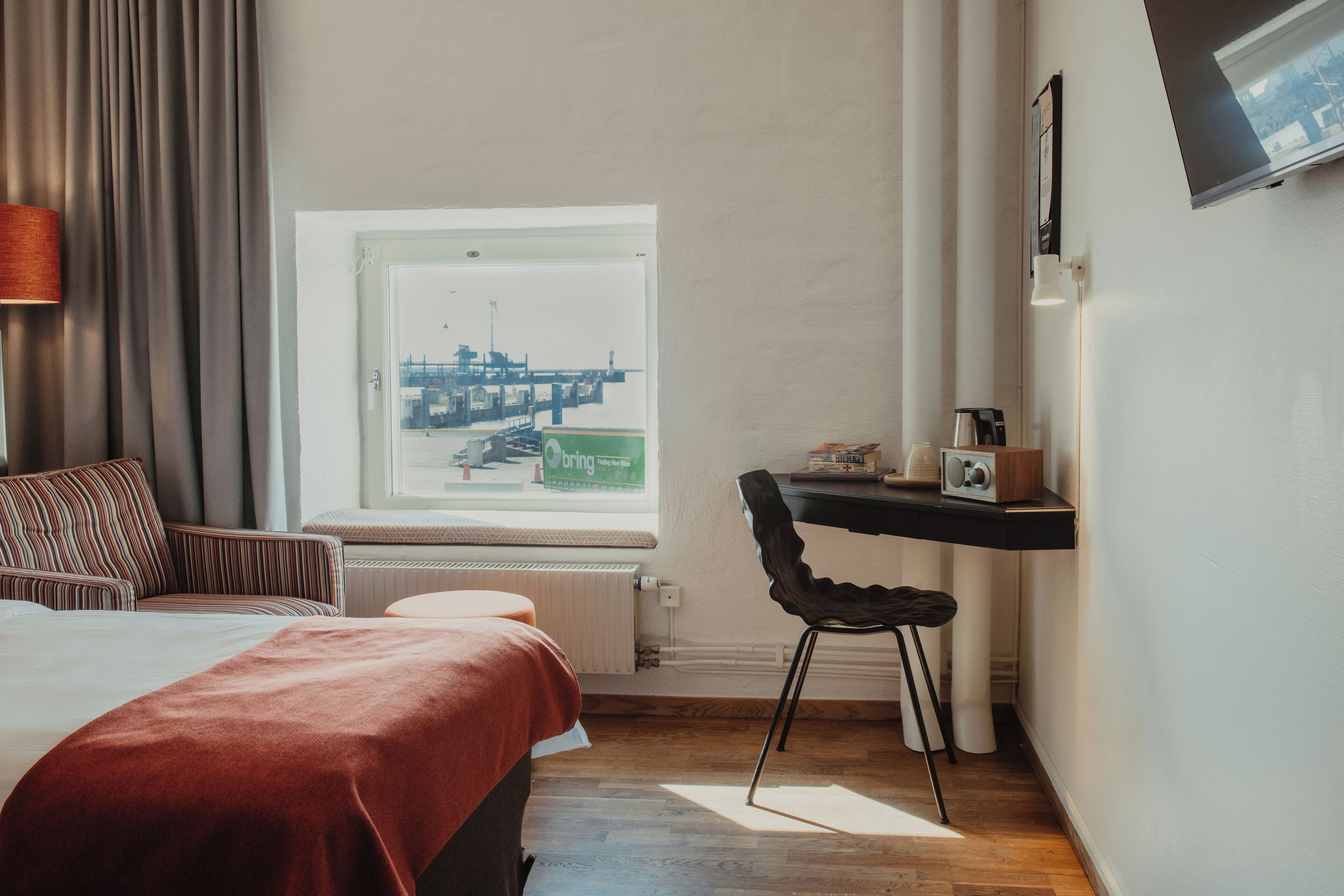 A hotel room with a bed, a window with a cityscape view, a desk with a chair, and a radiator.