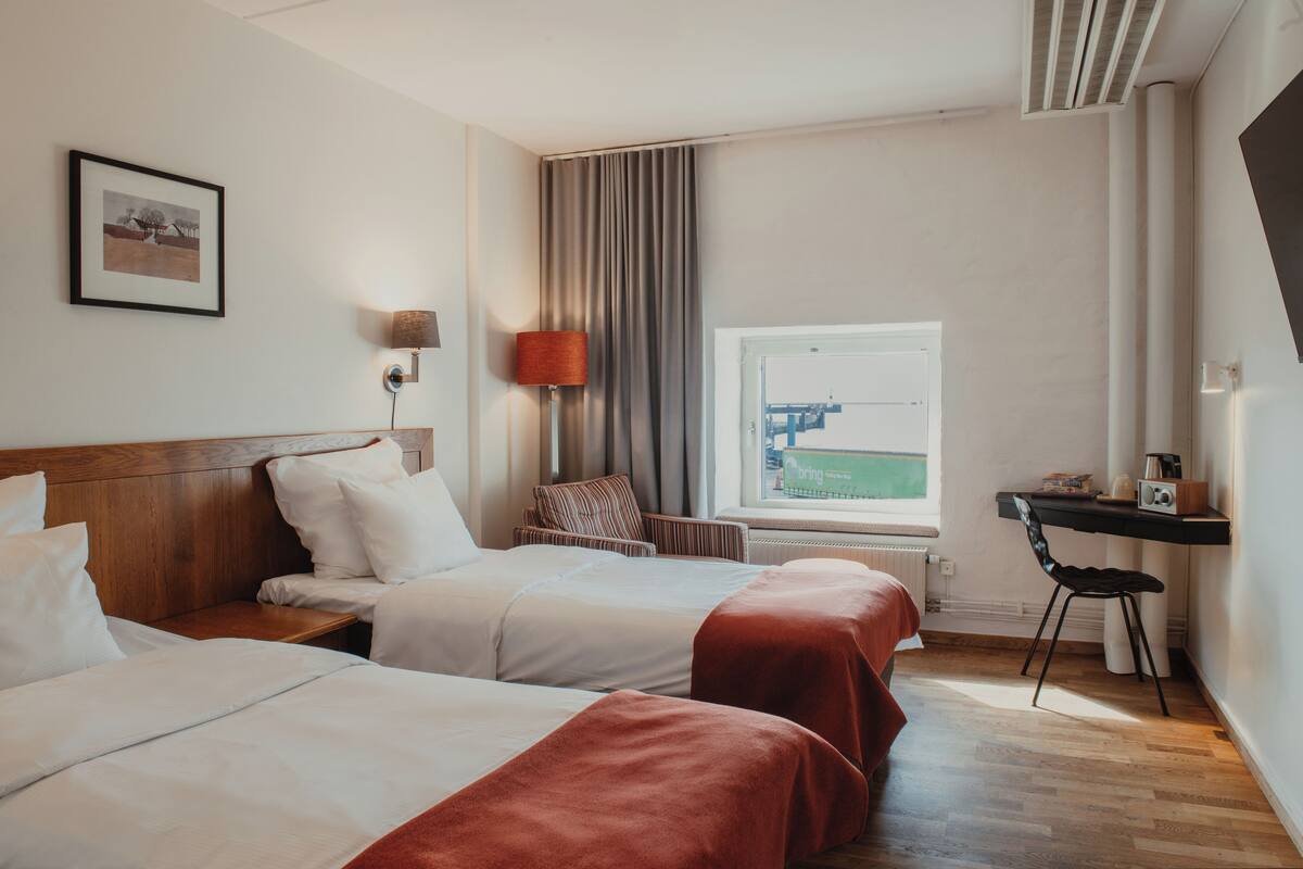 Una habitación de hotel con dos camas, un escritorio, una silla, una lámpara, una ventana con vistas y un cuadro en la pared.