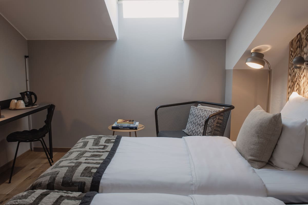 Una habitación pequeña con una cama, un escritorio y una silla. Hay un tragaluz sobre la cama y una lámpara en el escritorio.
