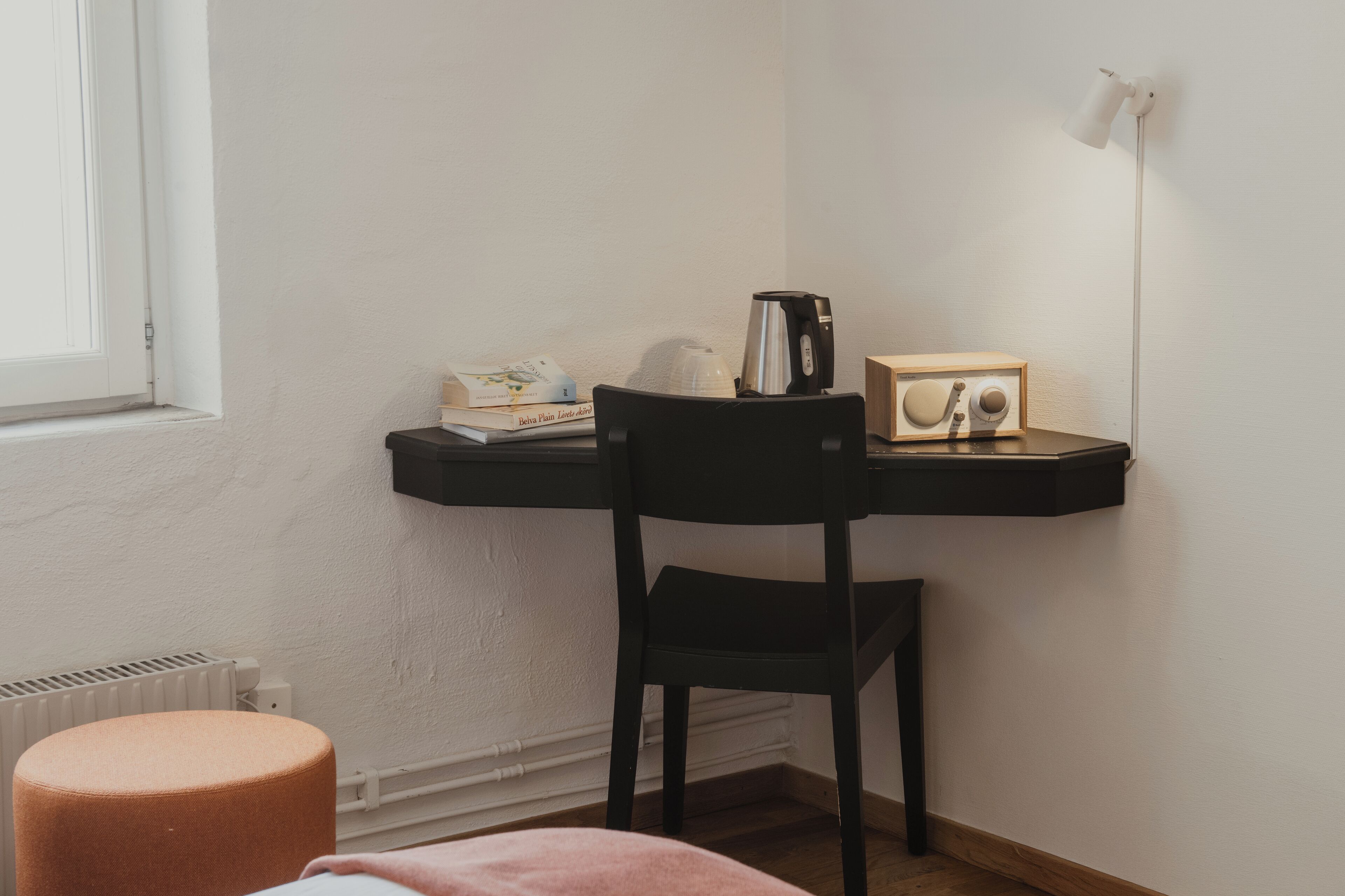 Una habitación pequeña y acogedora con una silla negra, un radiador blanco, un taburete de madera, una estantería con libros y un hervidor, además de una lámpara de pared.