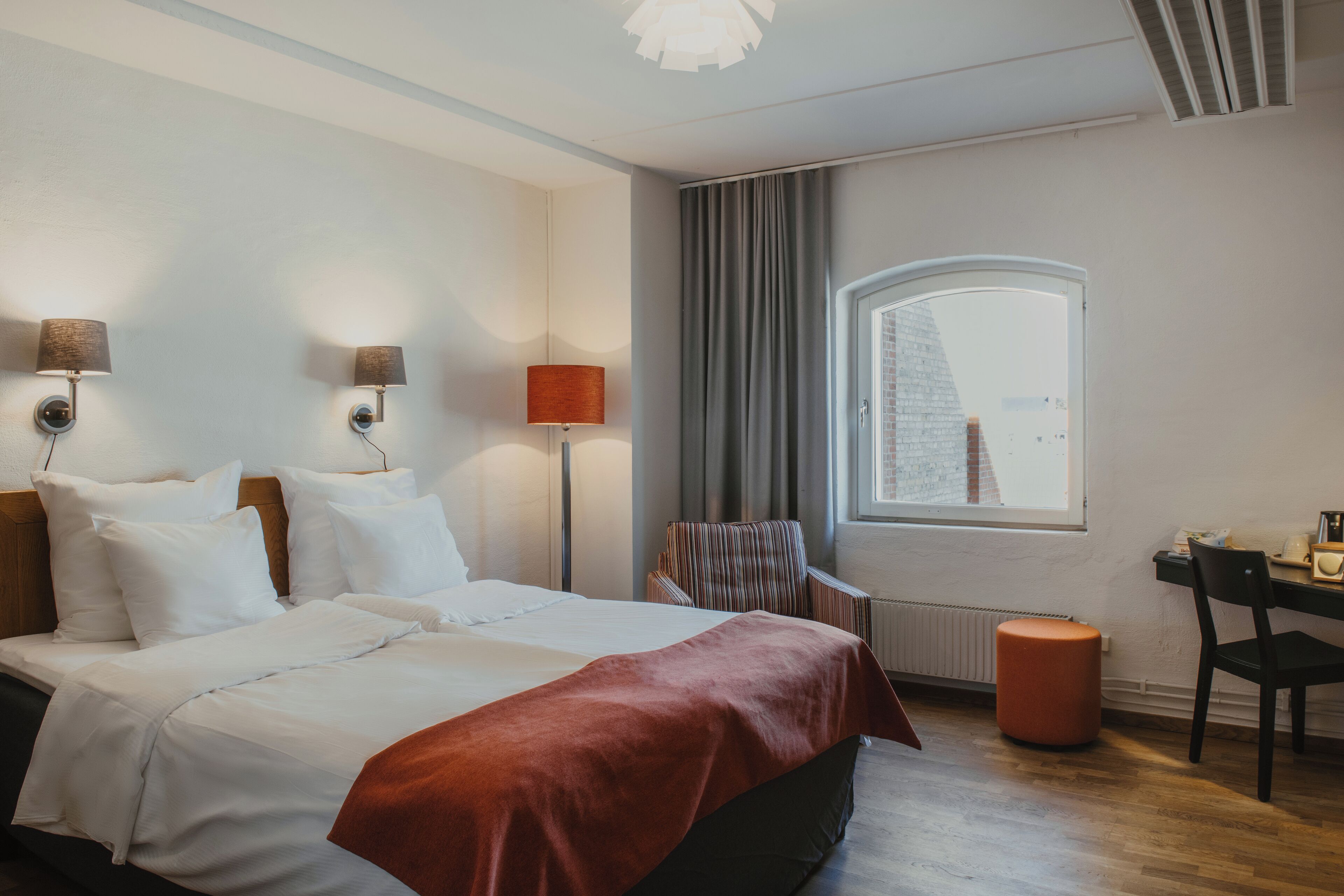 Ein Hotelzimmer mit einem großen Bett, zwei Nachttischlampen, einem Schreibtisch mit Stuhl, einem Fenster mit Vorhängen und einem Heizkörper.