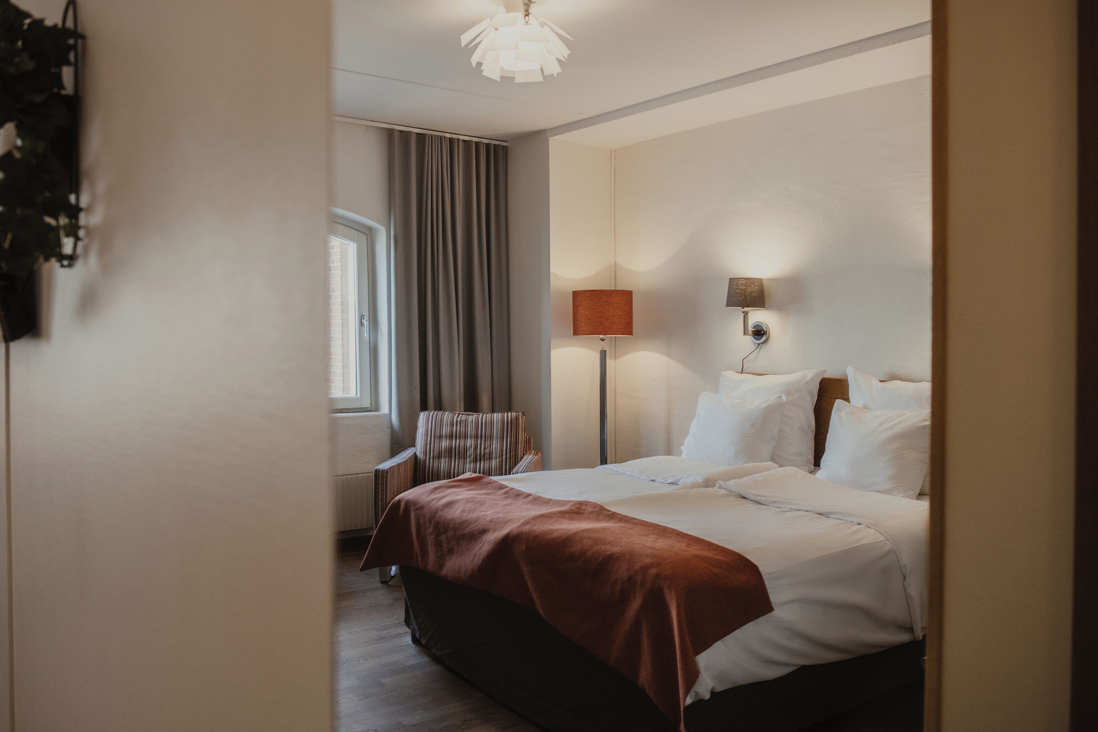 Une chambre d’hôtel avec un lit, une fenêtre avec des rideaux, une lampe et un luminaire fixé au mur.