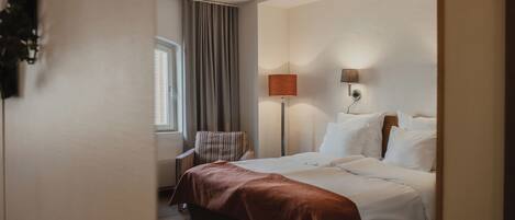 Une chambre d’hôtel avec un lit, une fenêtre avec des rideaux, une lampe et un luminaire fixé au mur.