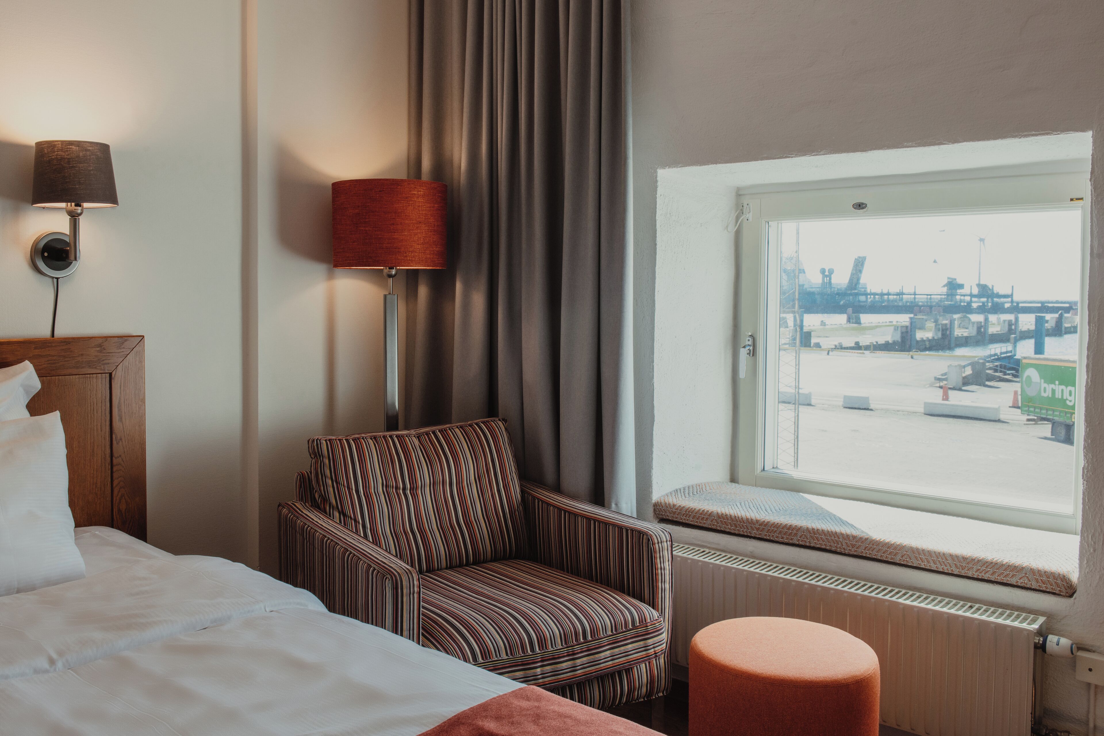 Une chambre d’hôtel avec un lit, un fauteuil à rayures, un pouf rouge, une fenêtre donnant sur un pont et une lampe fixée au mur.