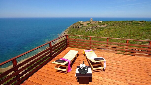 Een houten terras met twee ligbedden en een tafel, met uitzicht op de kust en een vuurtoren in de verte.