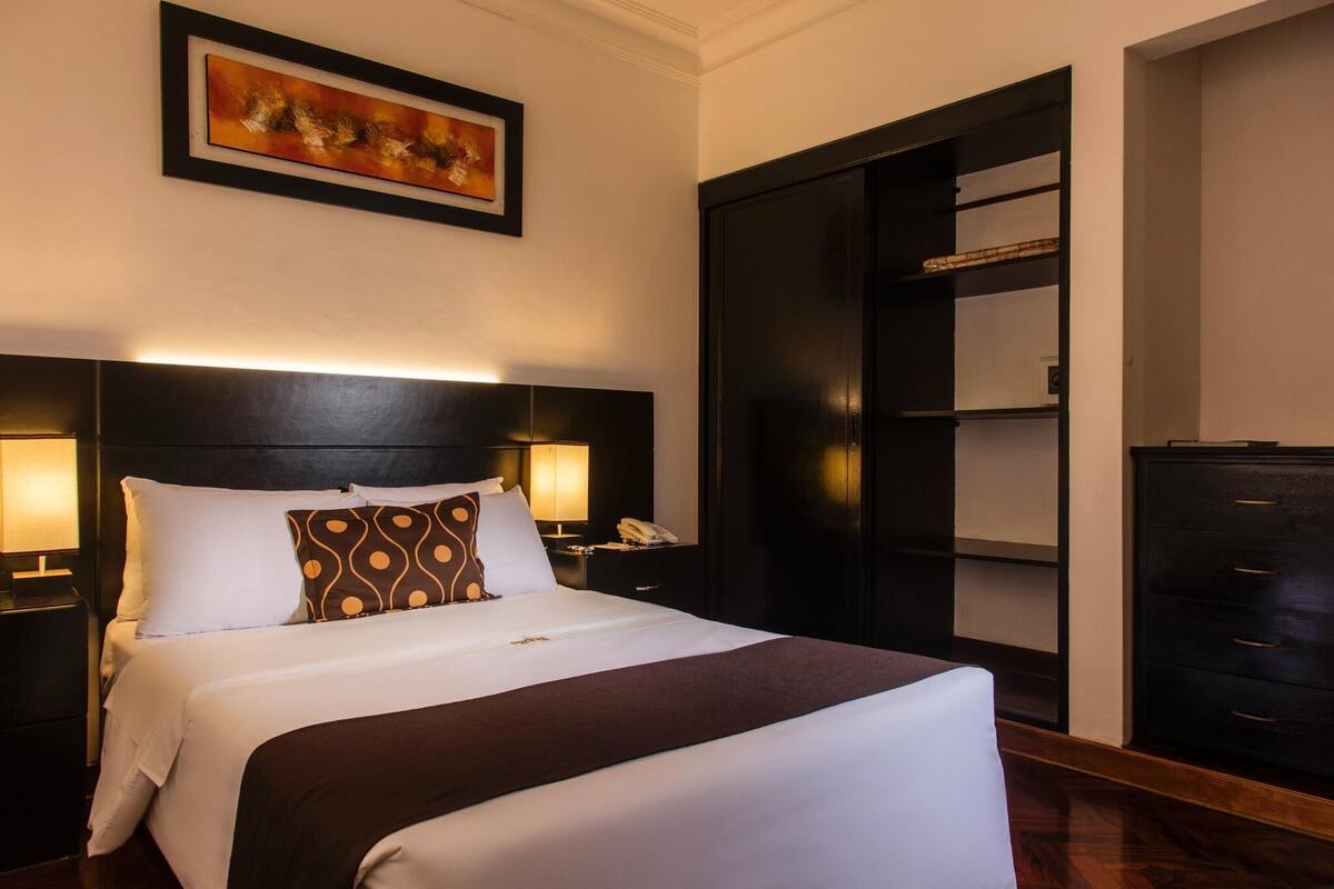 Habitación de hotel con una cama grande, lámparas de noche, mesita de noche, armario y un cuadro en la pared.