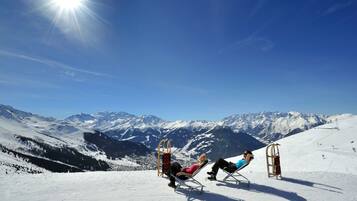 Due persone si rilassano su sdraio su un pendio montuoso innevato, con vista chiara su montagne innevate sullo sfondo.