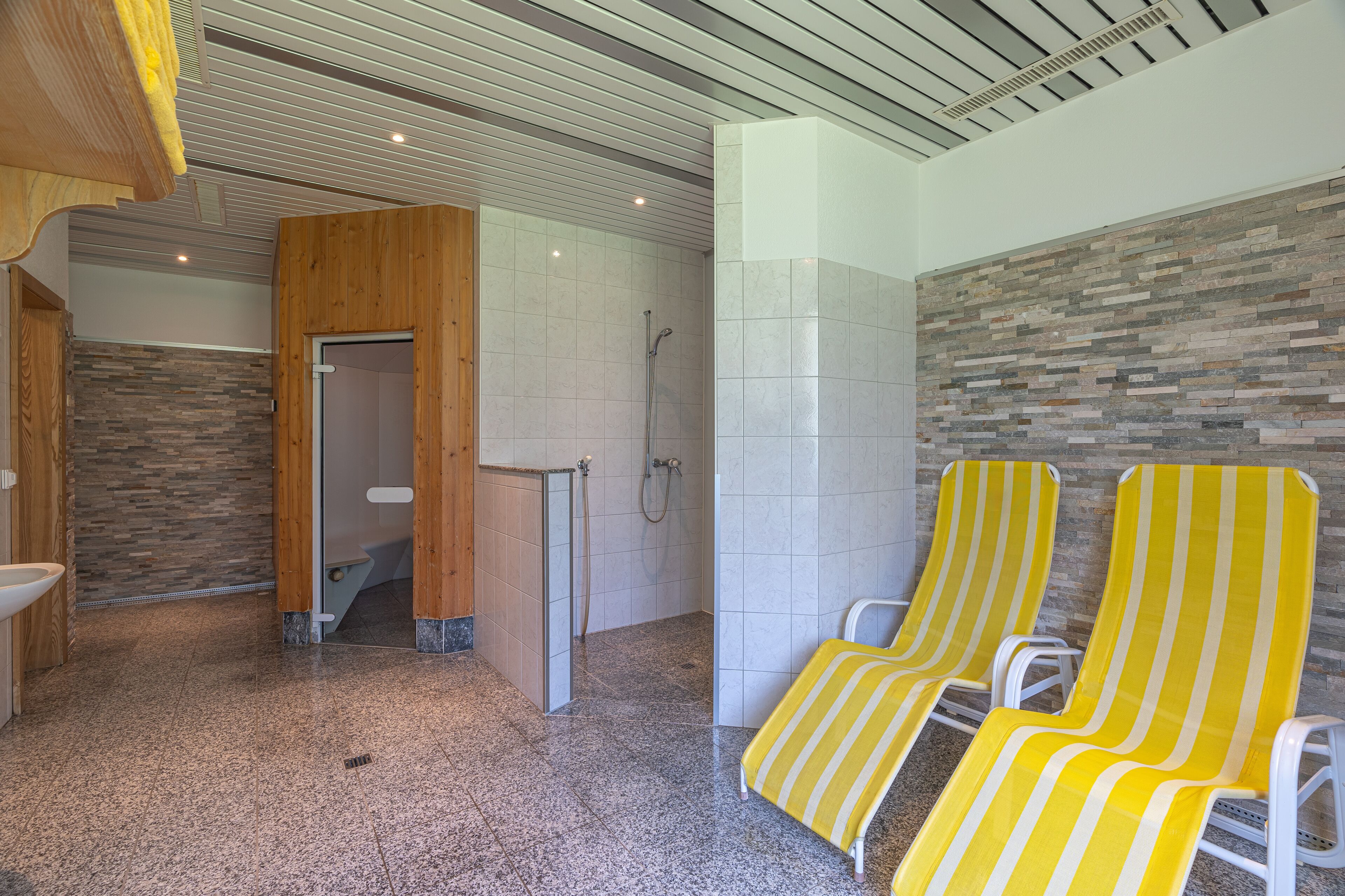 A modern bathroom with a shower, two yellow-striped lounge chairs, and a stone wall.