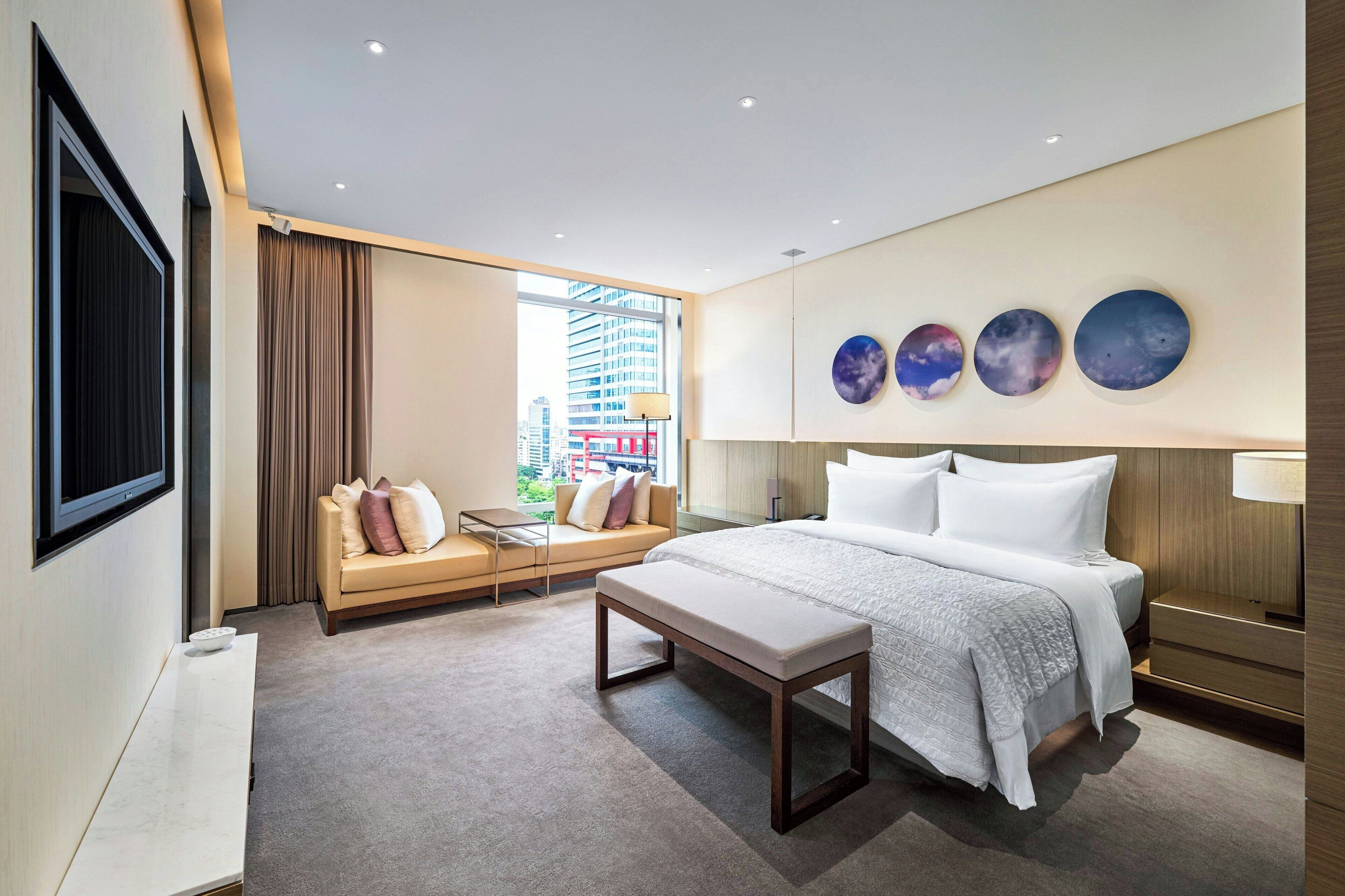 A modern hotel room with a large bed, a sofa, a bench, and a TV.