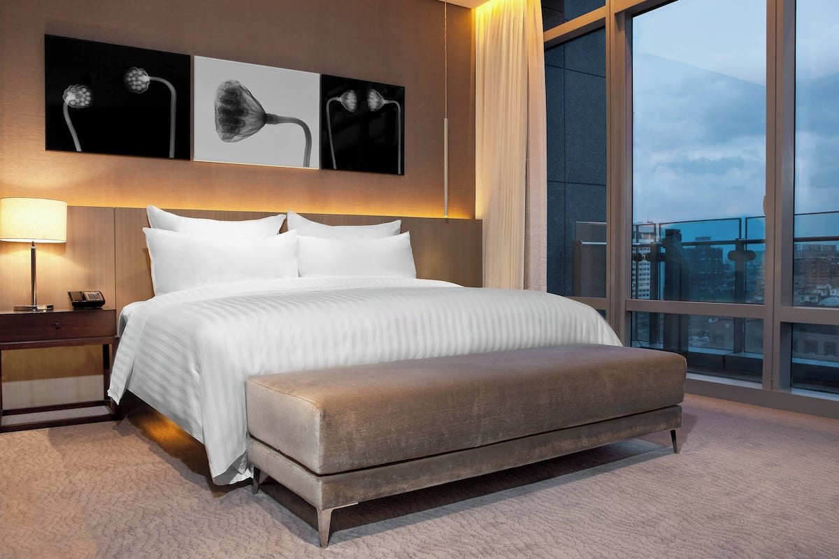 A modern hotel room with a large bed, a bedside table with a lamp, and a view of the city through large windows.