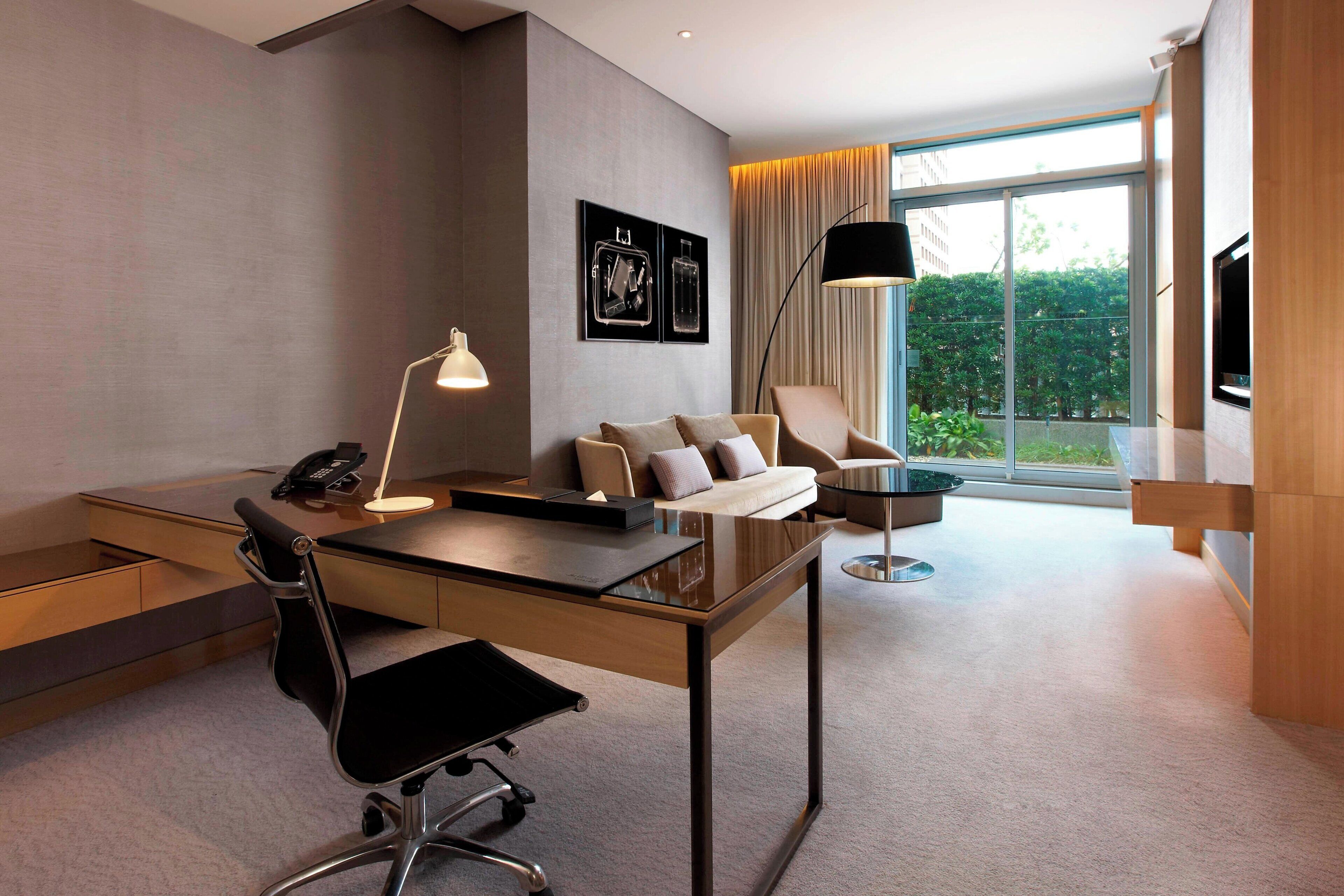 A modern hotel room with a desk, chair, sofa, and a large window offering a view of the outdoors.