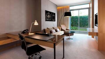 A modern hotel room with a desk, chair, sofa, and a large window offering a view of the outdoors.