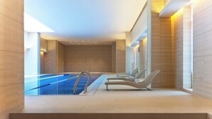 Uma piscina interior moderna com água azul cristalina, rodeada por paredes revestidas a azulejos bege e degraus que dão acesso à área da piscina.