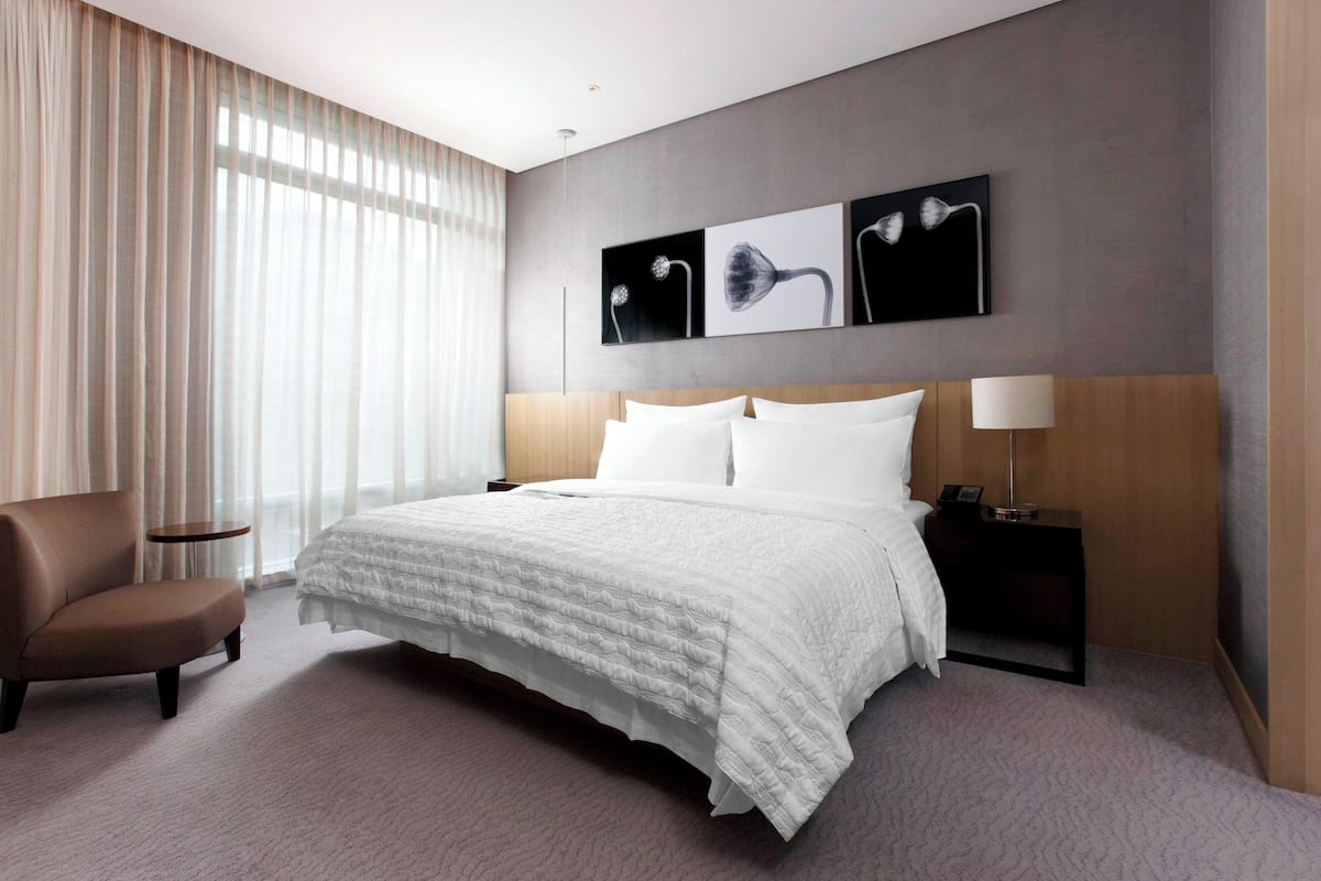 A modern hotel room with a large bed, a chair, a nightstand, and a wall-mounted lamp.