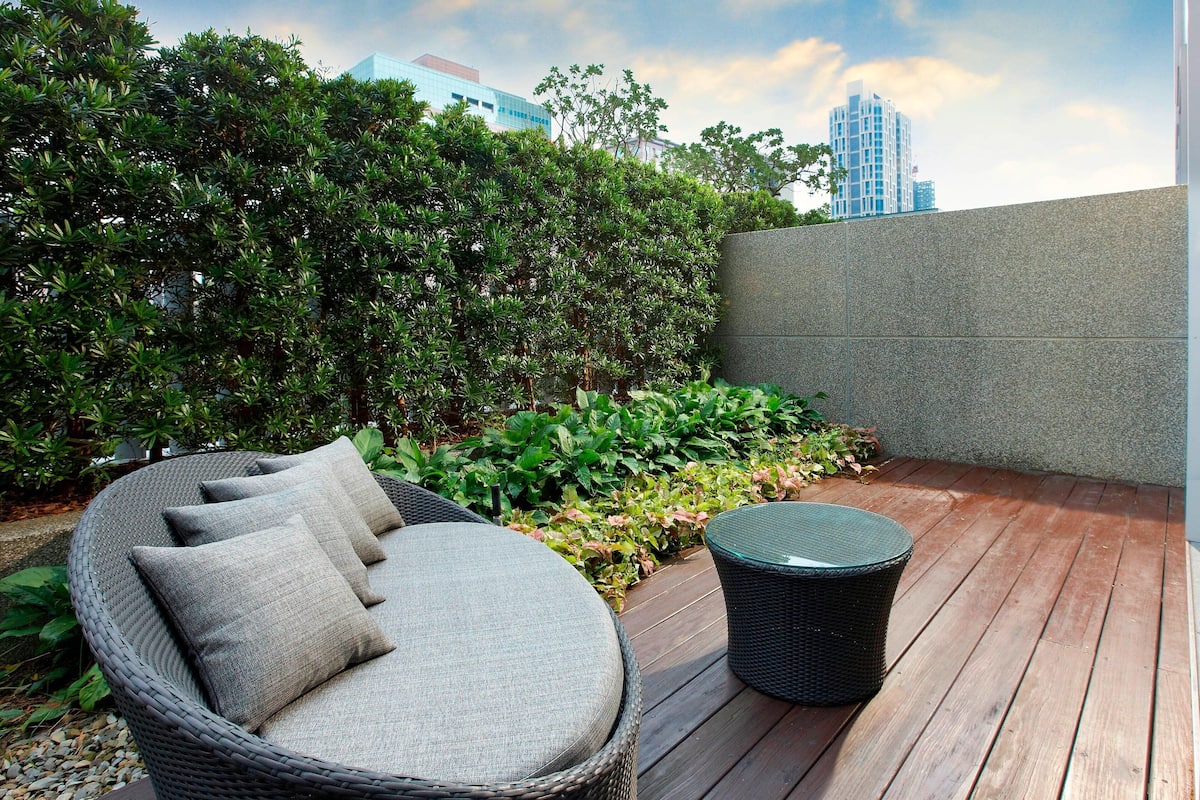 A wicker lounge chair with cushions, a round table, and a wooden deck with a low wall and greenery.