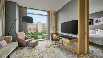 A hotel room with a large window, a desk, a sofa, a TV, and a bed.