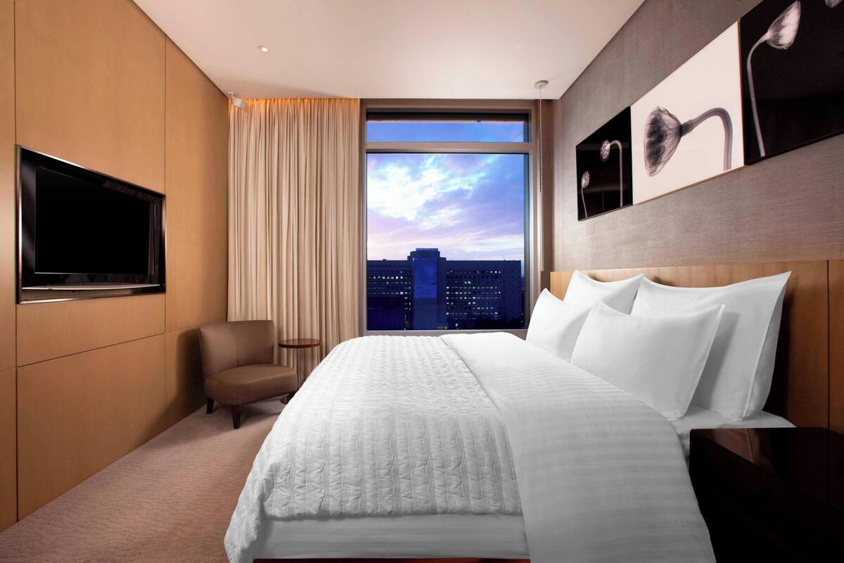 A hotel room with a large bed, a TV mounted on the wall, a chair, and a window with a cityscape view.