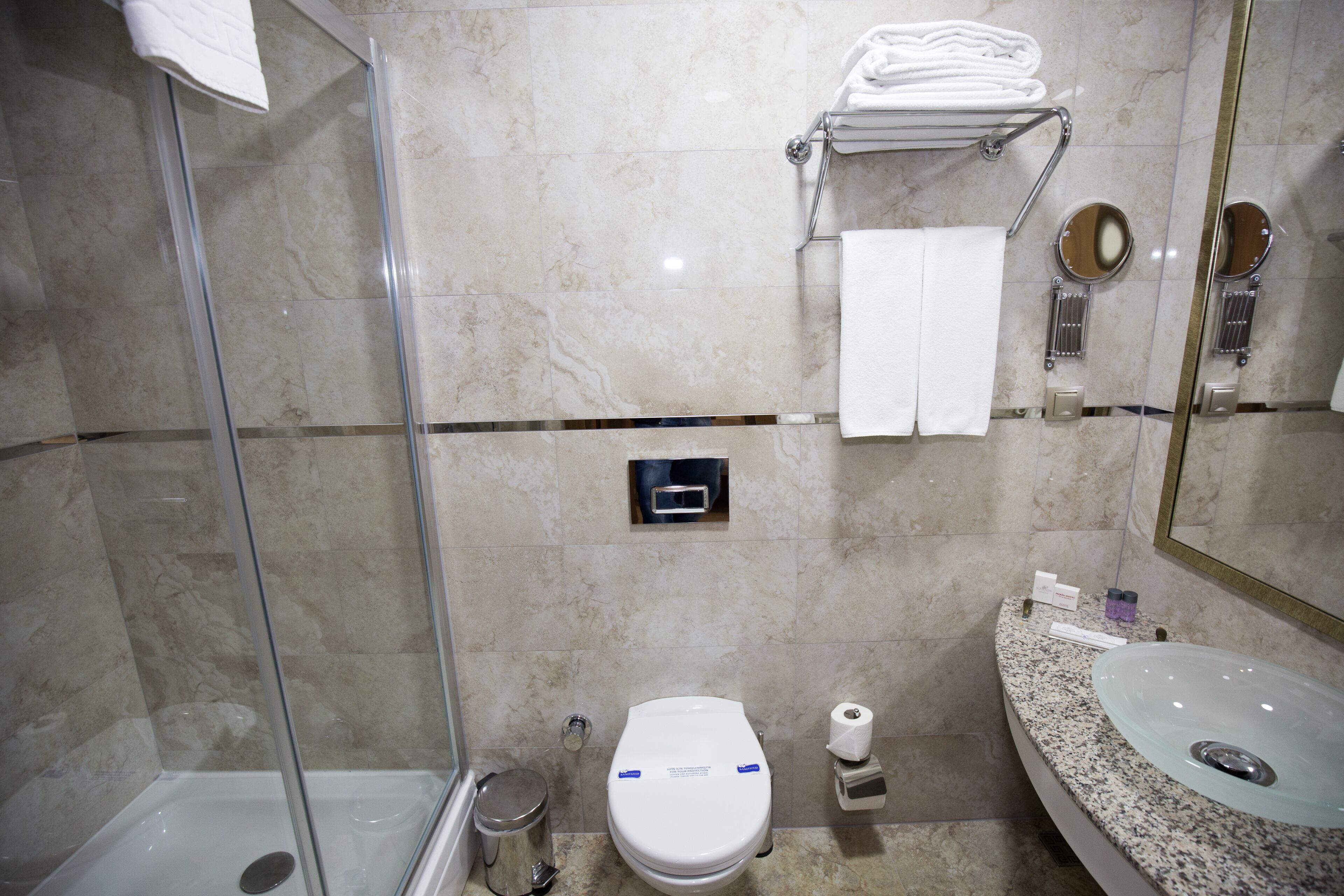 A bathroom with a glass shower enclosure, a toilet, a sink, and a towel rack with folded towels.