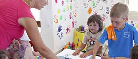 A classroom with children engaged in painting activities, a teacher assisting them, and colorful handprints on the wall.