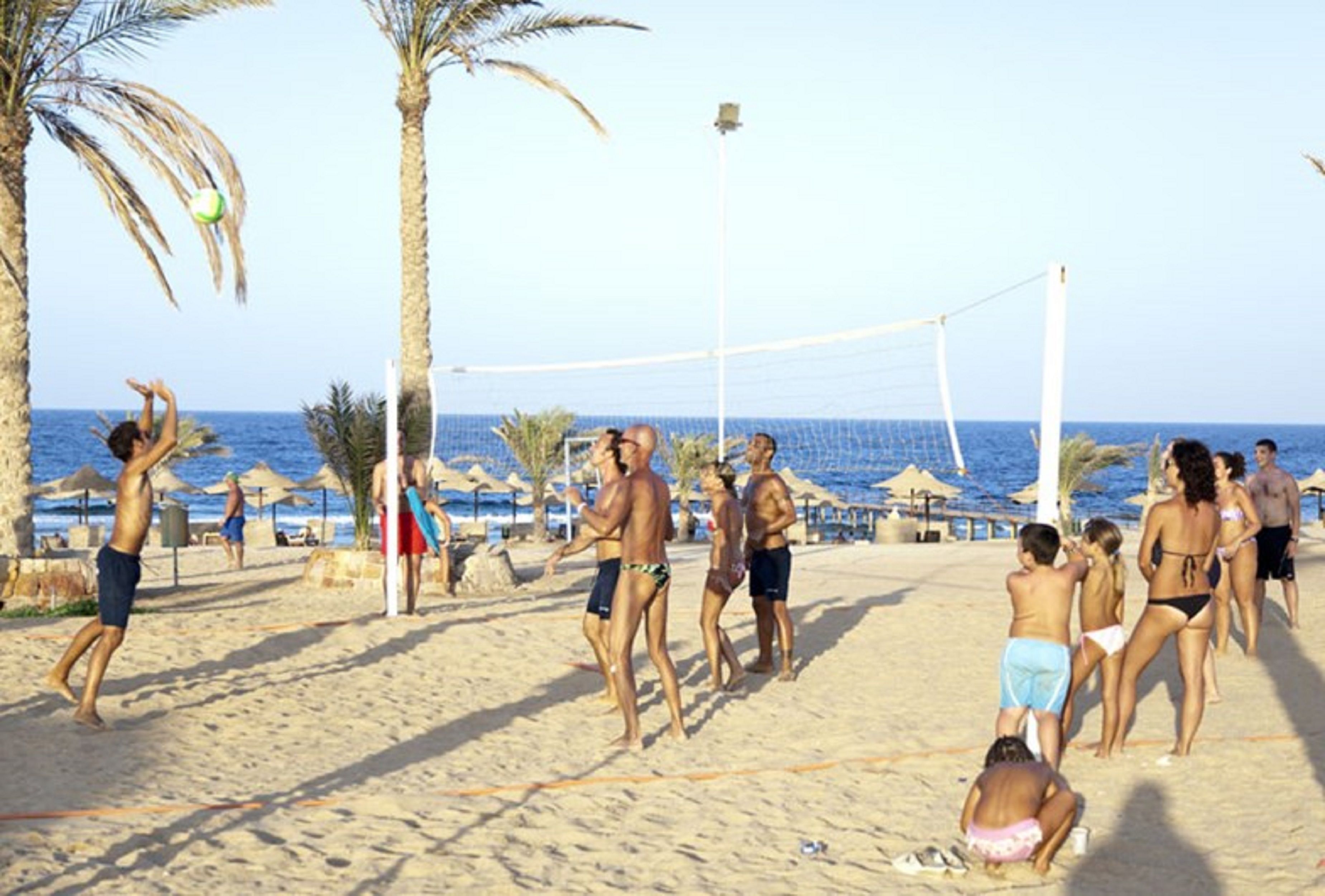 Volleyballkamp på stranden med spillere og tilskuere på sandbanen, palmer og havet i baggrunden.