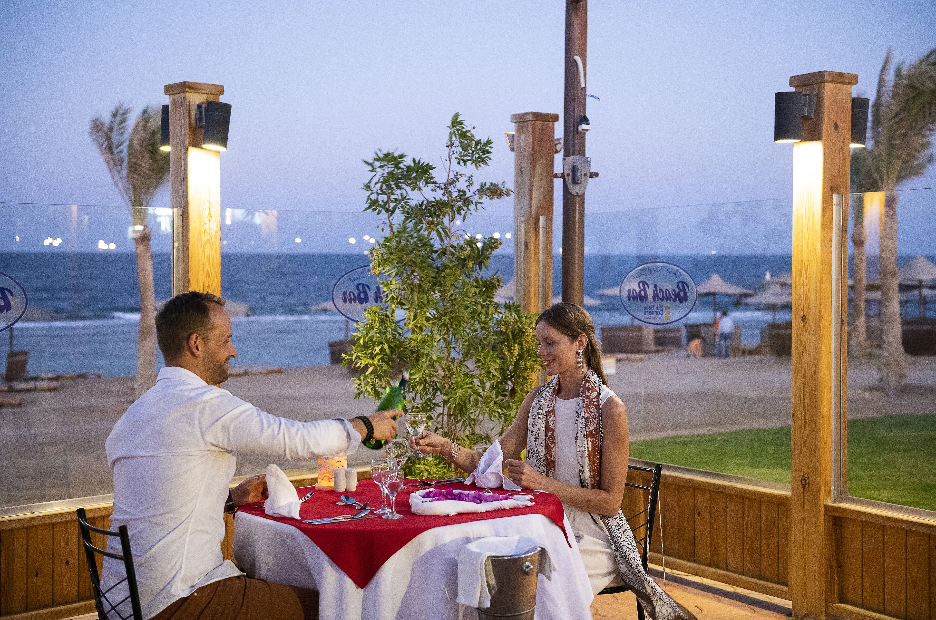 A couple dining at a wooden table with a red tablecloth, overlooking the sea.