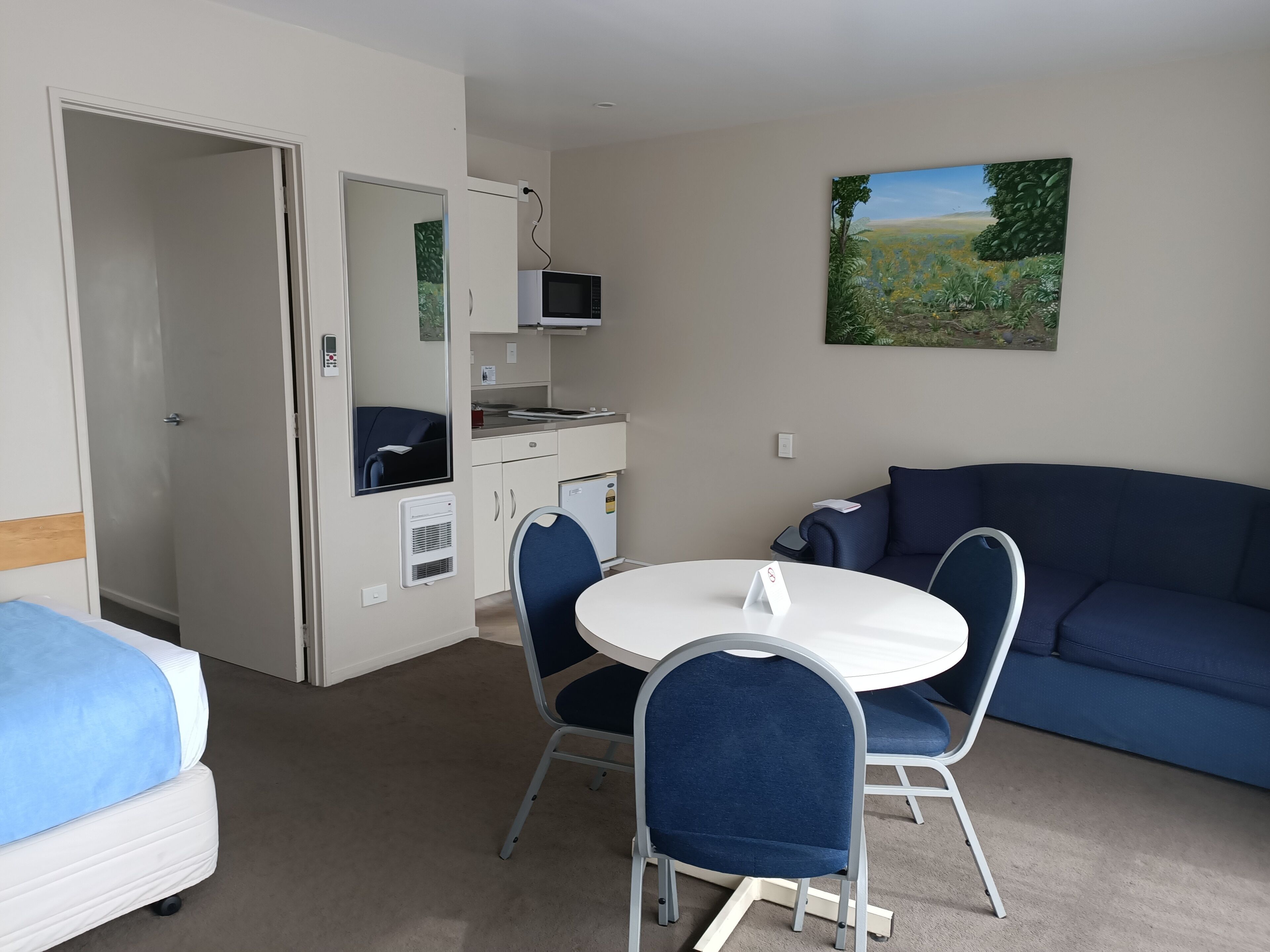 A compact room with a bed, a dining table with chairs, a kitchenette, and a blue sofa.