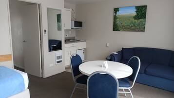 A compact room with a bed, a dining table with chairs, a kitchenette, and a blue sofa.