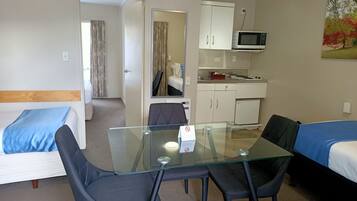 A hotel room with a glass table, two chairs, a bed with blue bedding, a kitchenette with a microwave, and a painting on the wall.