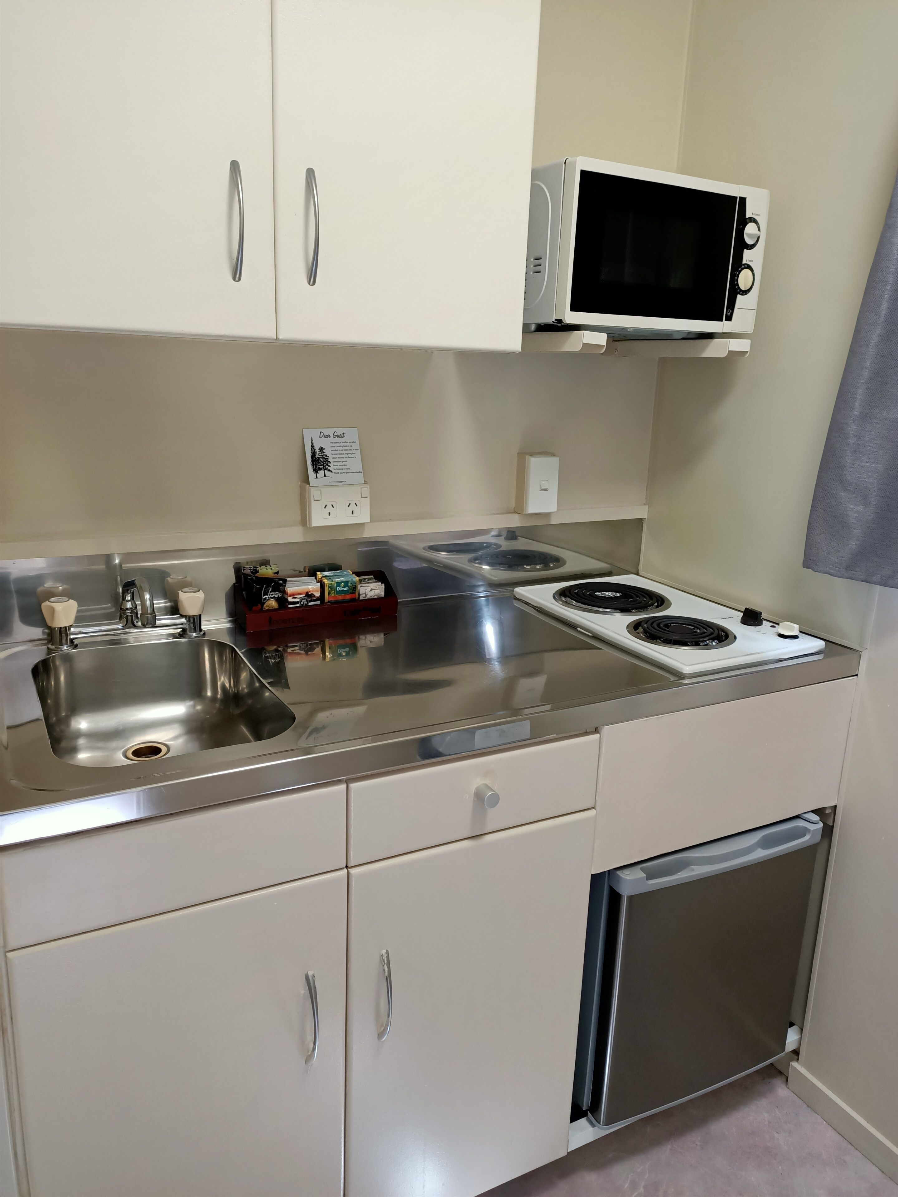 A compact kitchen with a stainless steel sink, a microwave, and a stove. There are cabinets with silver handles and a small refrigerator below the counter.