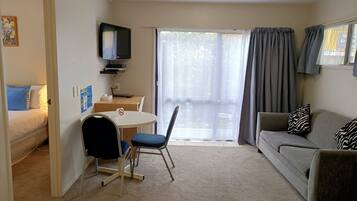 A compact living space with a bed, a sofa, a dining table with chairs, a TV mounted on the wall, and a sliding glass door leading to an outdoor area.