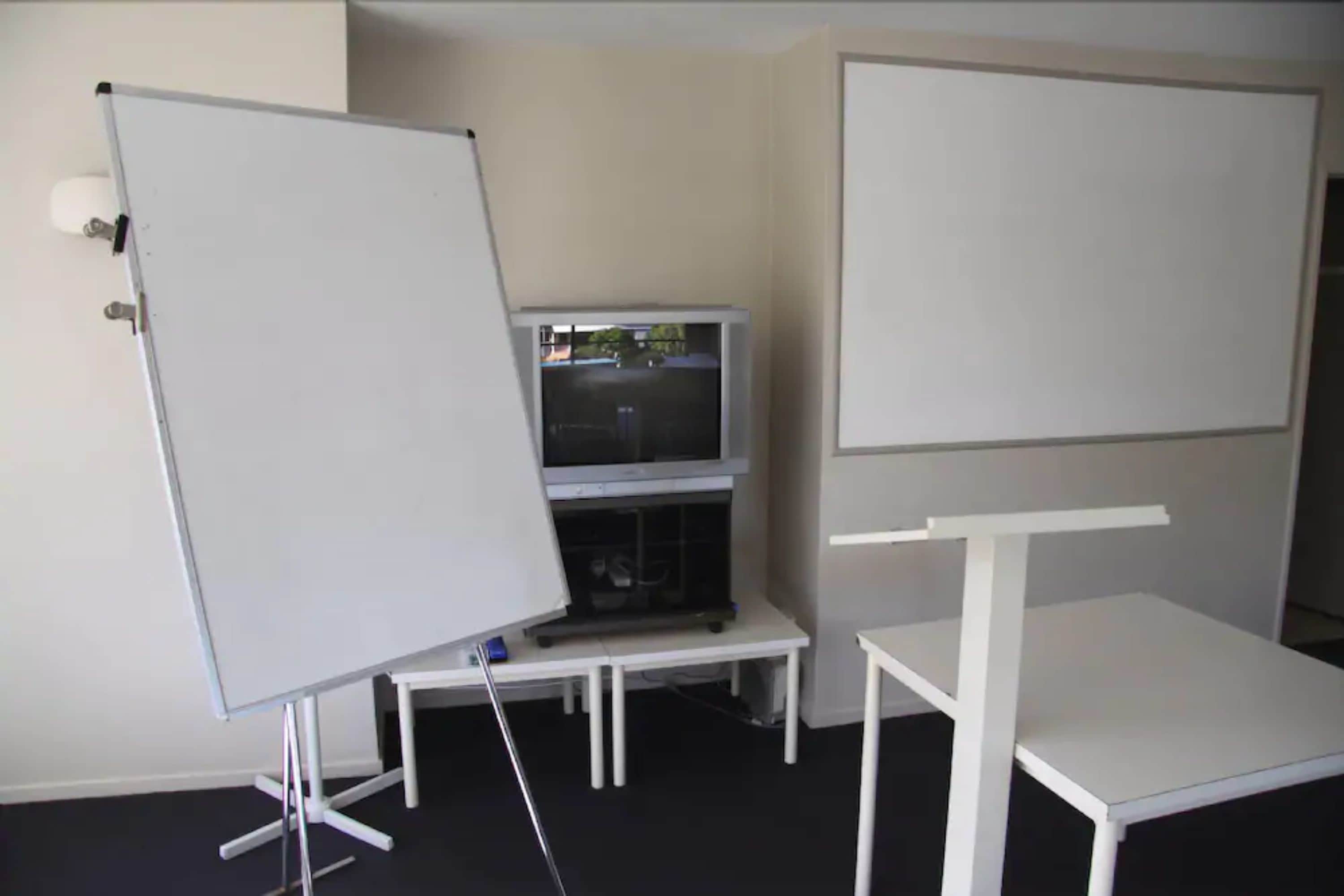 A room with a whiteboard, a television on a stand, and a white table.