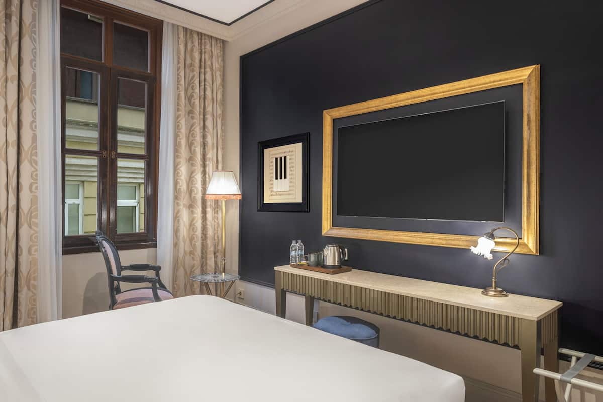 A hotel room with a bed, a desk with a lamp, a chair, a window with curtains, and a framed picture on the wall.