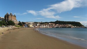 A wide sandy beach with a historic building on the left and a hill with a fort on the right. - Palm Court Hotel (Scarborough)