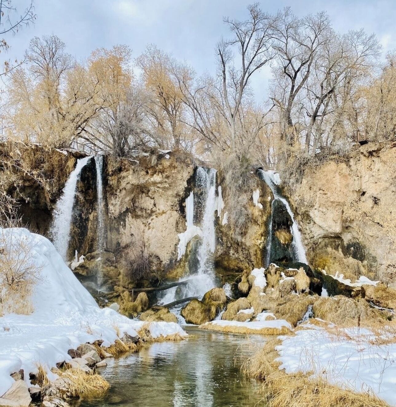 Una cascada natural con agua cristalina que fluye sobre rocas, rodeada de un paisaje nevado y árboles sin hojas.