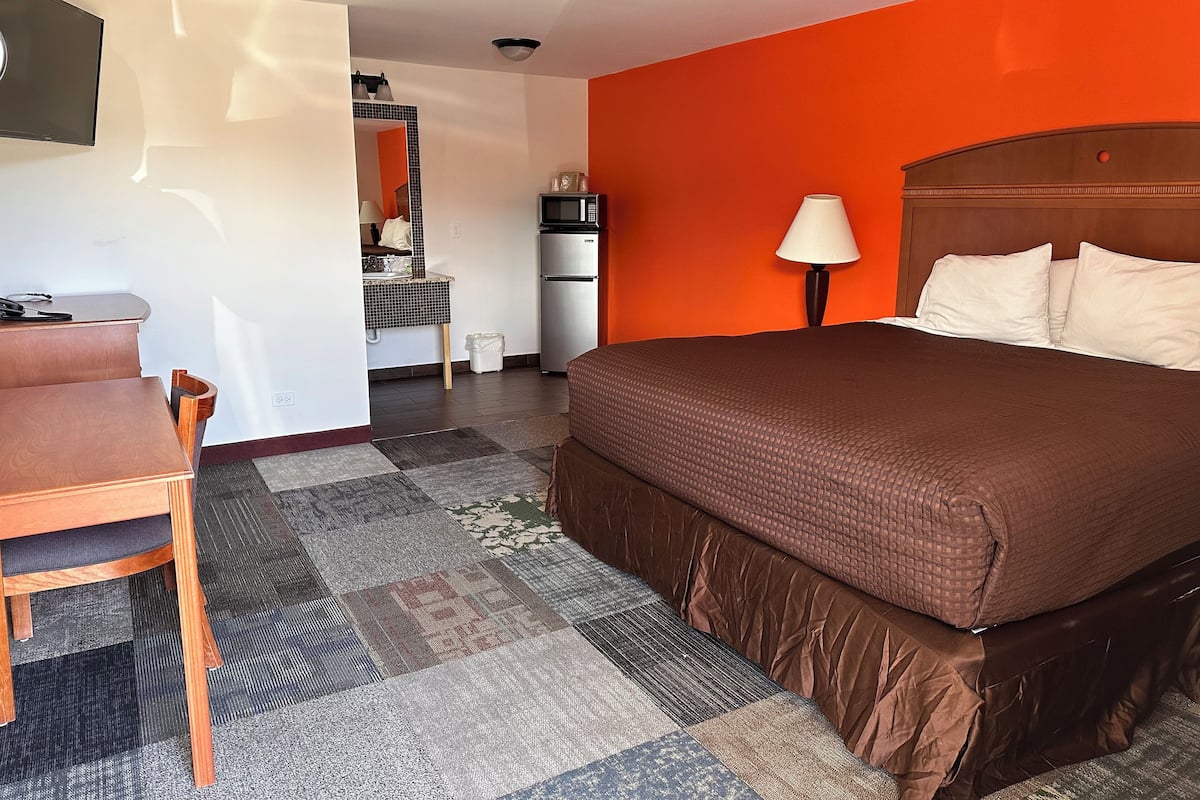 Habitación de hotel con cama, escritorio, silla, televisión, lámpara y kitchenette.